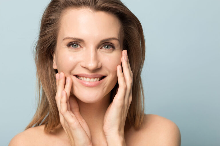 Woman with healthy, glowing skin smiling and touching her face to illustrate RF microneedling vs microneedling results.