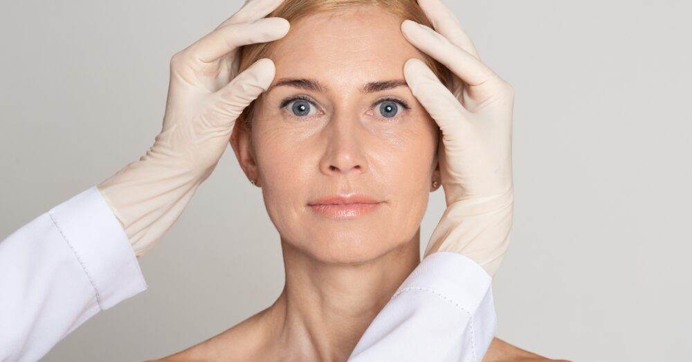 Gloved hands examining a woman's forehead and temple area before a Botox treatment