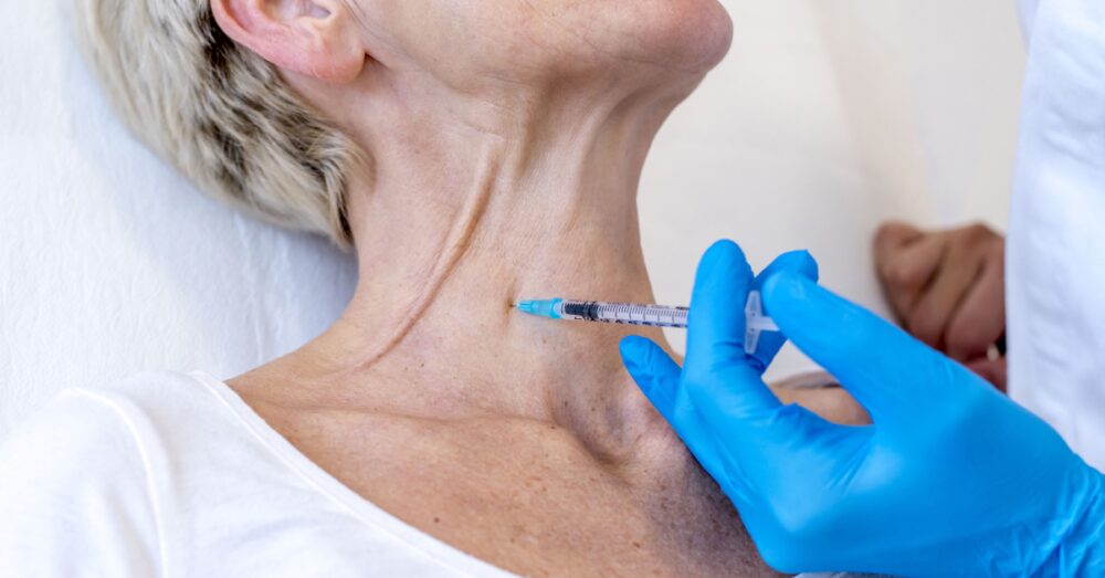A gloved hand holding a syringe near the neck area of a woman during an injectable treatment procedure
