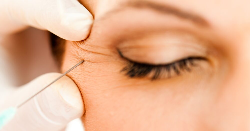 A close-up of a woman's face with her eyes closed as a gloved hand prepares to administer a cosmetic injection near her eye area