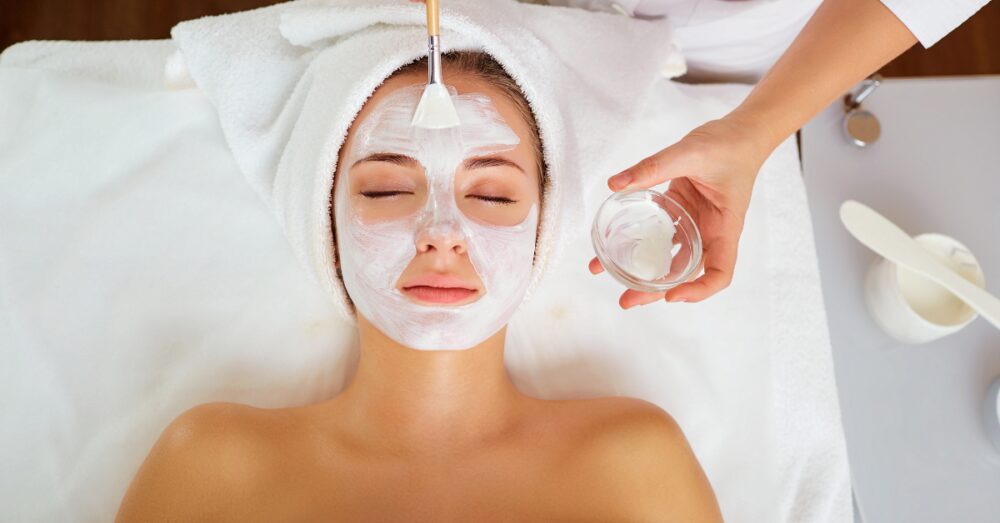 A woman lying down while an aesthetician applies a white facial mask with a brush during a professional facial treatment