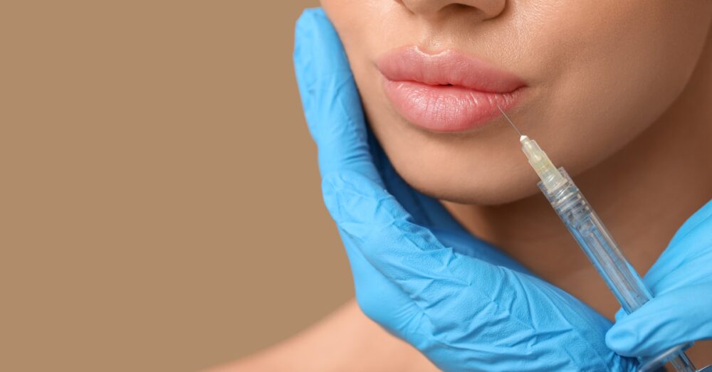 Close-up of a woman's lower face receiving a lip filler injection administered by a gloved hand holding a syringe