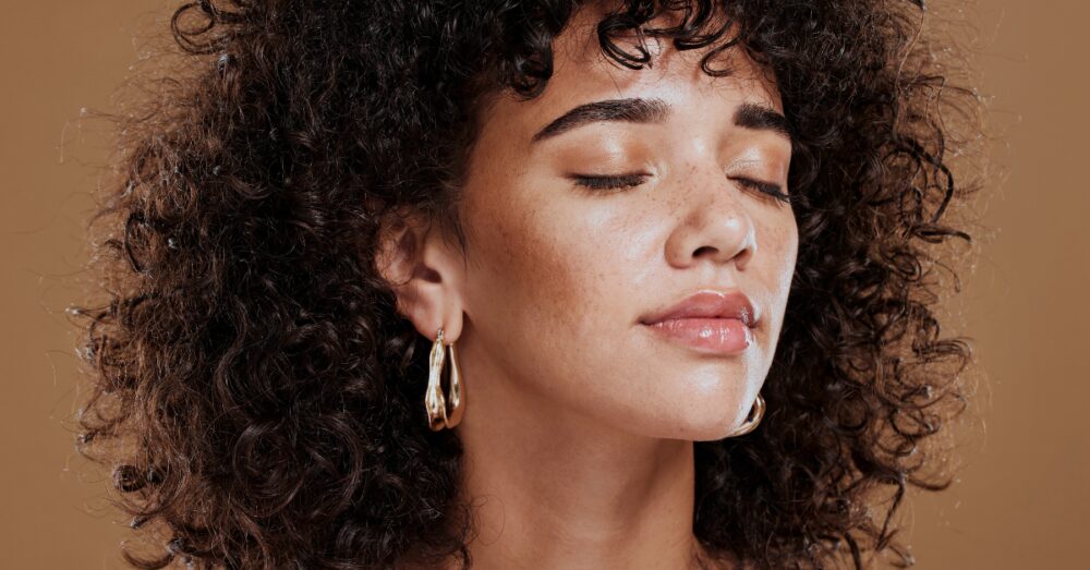 A young woman with curly hair and gold hoop earrings posing with eyes closed against a warm brown background