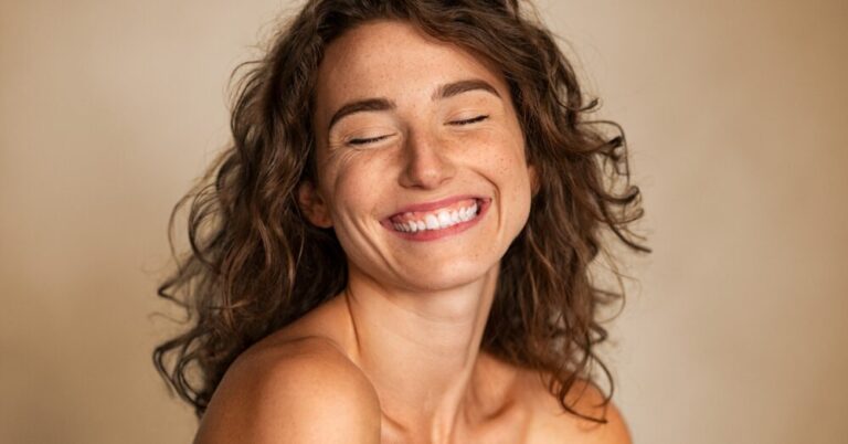 Close up of a woman with freckles and curly hair smiling with her eyes closed