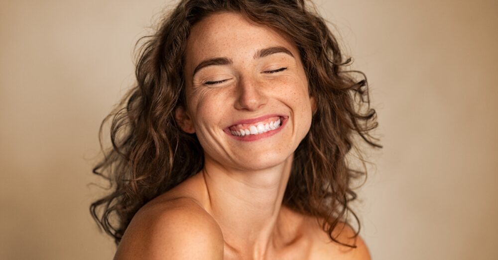 Close up of a woman with freckles and curly hair smiling with her eyes closed